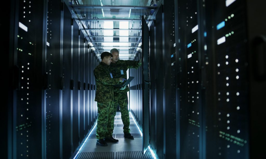 In Data Center Two Military Men Work with Open Server Rack Cabinet. One Holds Military Edition Laptop.